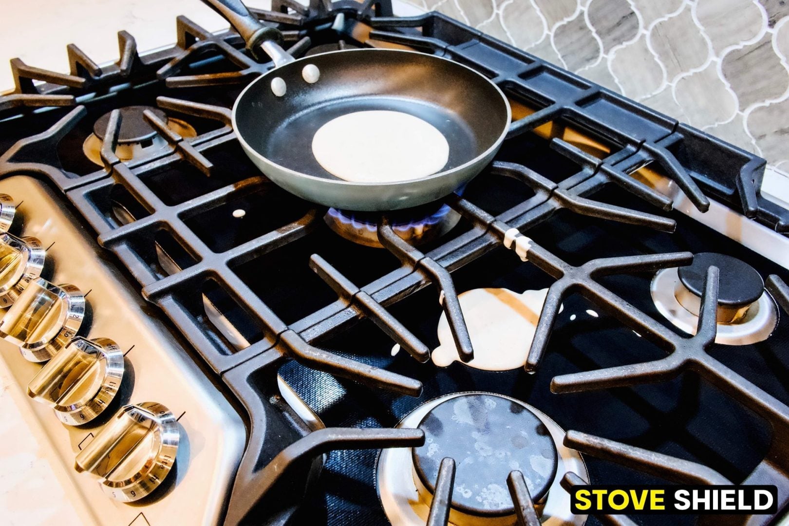 A frying pan with a tortilla is on a lit gas stove. A protective cover labeled STOVE SHIELD is installed on the stove grates.