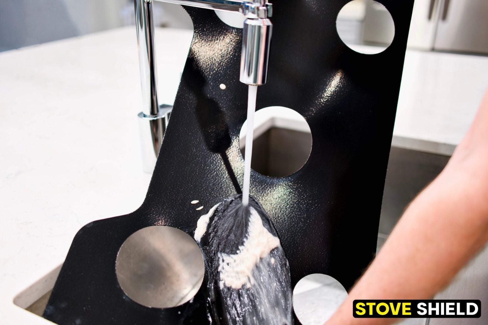 Hand washing a black metal stove shield with circular cutouts under running water, with STOVE SHIELD branding in the corner